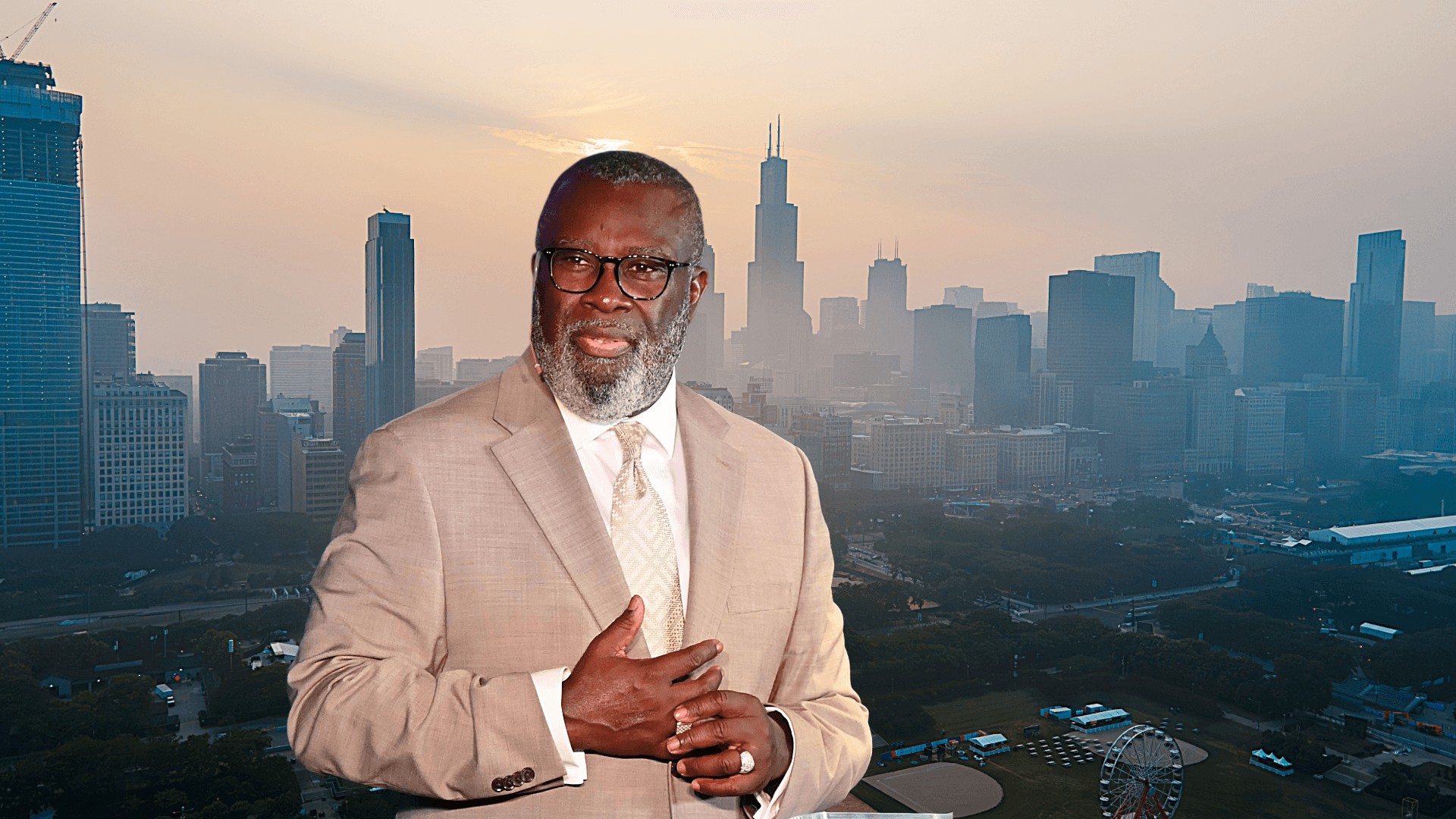 Man in a tan suit and glasses gesturing against a hazy Chicago skyline at sunset.